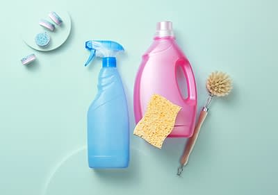 Eine blaue Sprühflasche und eine pinke Waschmittelflasche stehen neben einem gelben Schwamm und einer Bürste mit Holzgriff auf einem hellblauen Hintergrund