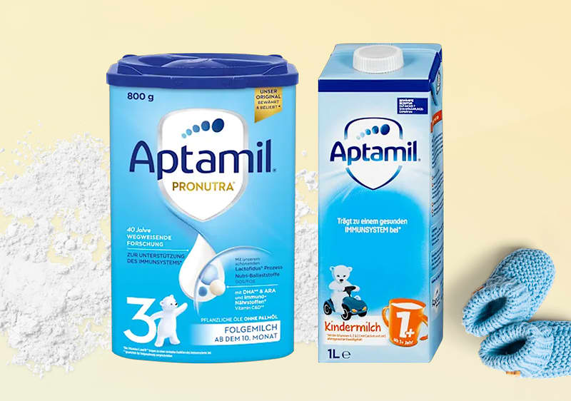 Eine blaue Aptamil Pronutra Folgemilch-Packung neben einer weißen Aptamil Kindermilch-Packung, daneben blaue Babyschuhe