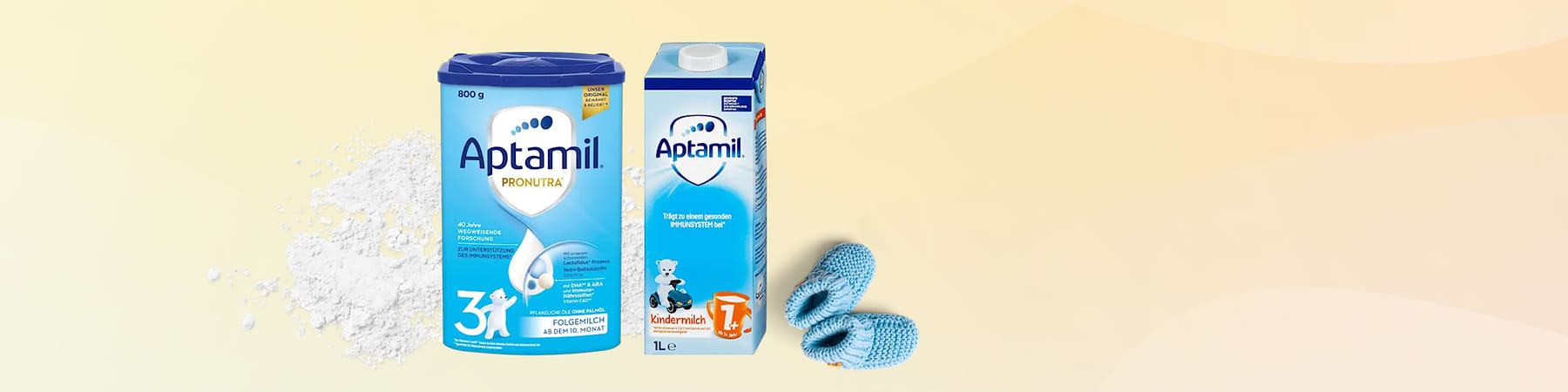 Eine blaue Aptamil Pronutra Folgemilch-Packung neben einer weißen Aptamil Kindermilch-Packung, daneben blaue Babyschuhe
