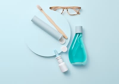 Eine Zahnbürste, eine Brille, eine blaue Zahnpastatube, eine blaue Mundspülungsflasche und ein Nasenspray auf hellblauem Hintergrund