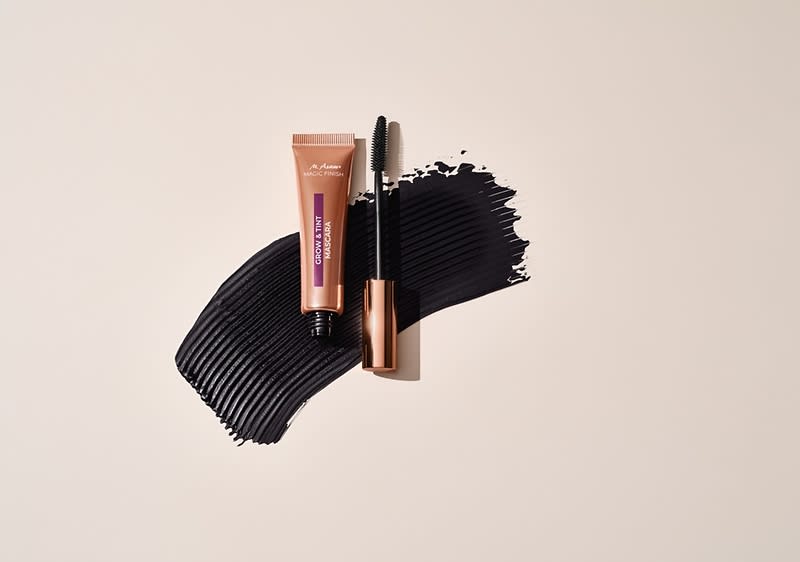 Eine braune Mascara-Tube mit lila Aufschrift „Volume Mascara“ neben einem geöffneten Mascara-Applikator auf schwarzem Farbstrich