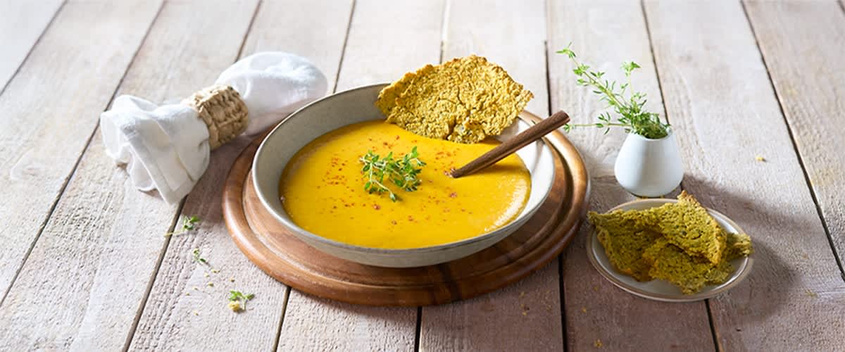 Eine Schale mit gelber Suppe, garniert mit frischen Kräutern, steht auf einem Holzbrett, daneben ein Stück knuspriges Brot