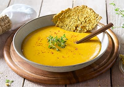 Eine Schüssel mit gelber Suppe, garniert mit Kräutern, einem Holzlöffel und einem Stück knusprigem Brot auf einem Holzbrett