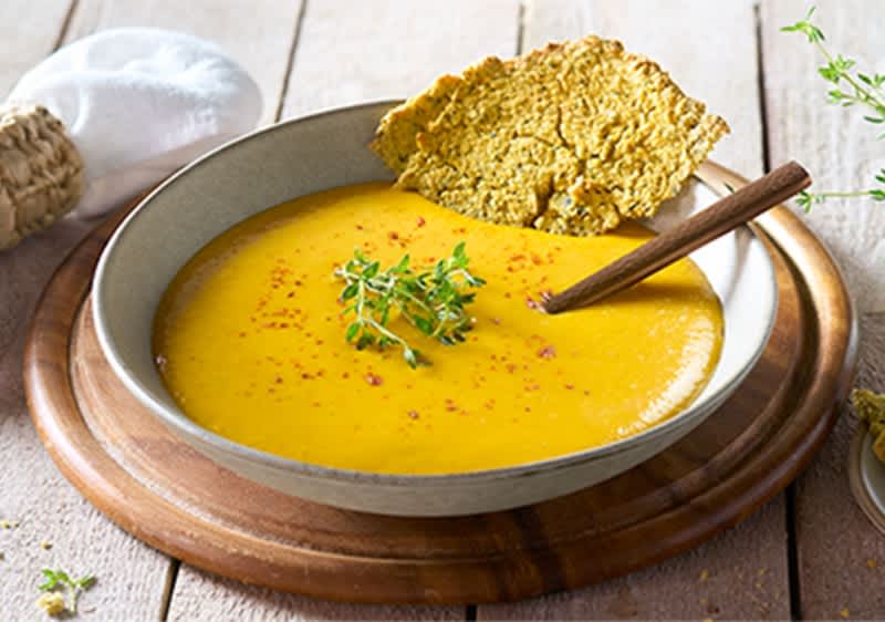 Eine Schüssel mit gelber Suppe, garniert mit Kräutern, einem Holzlöffel und einem Stück knusprigem Brot auf einem Holzbrett