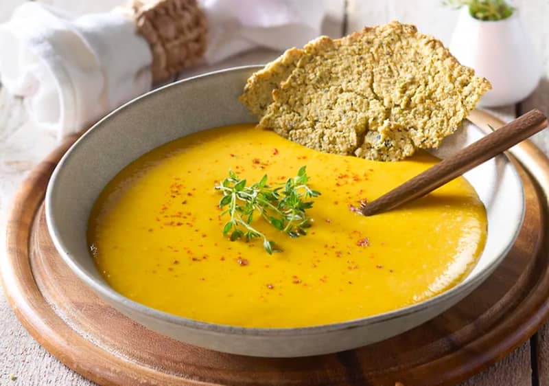 Eine Schüssel mit gelber Suppe, garniert mit frischem Thymian, einem Holzlöffel und einem Stück knusprigem Brot auf einem Holzbrett