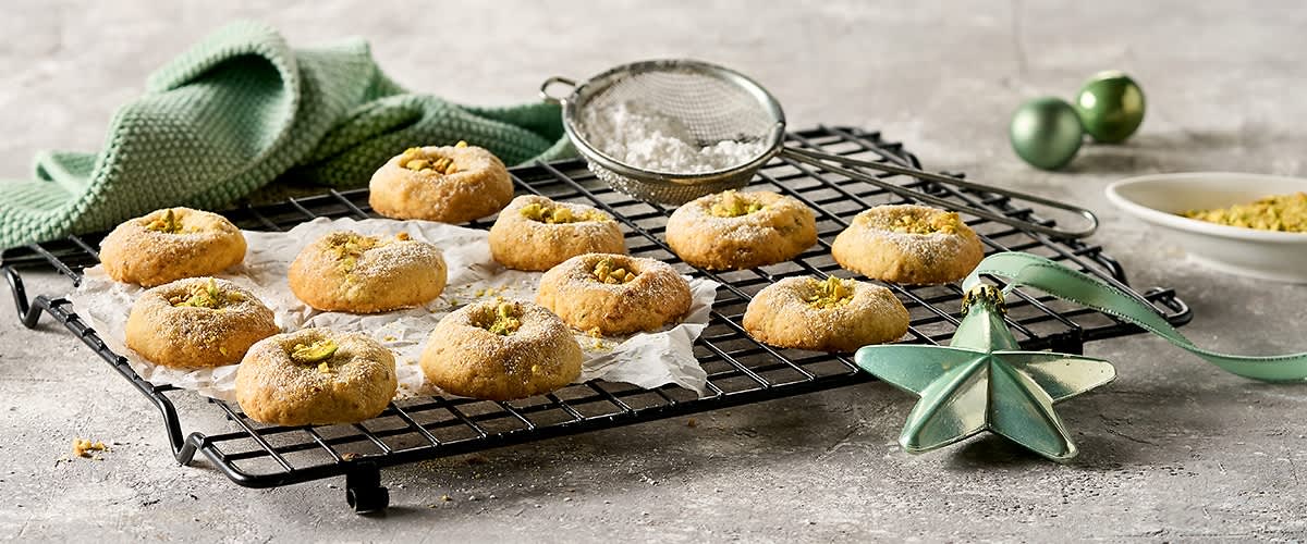Mehrere Kekse mit Pistazien auf einem Gitterrost, daneben ein grünes Tuch und ein Sieb mit Puderzucker