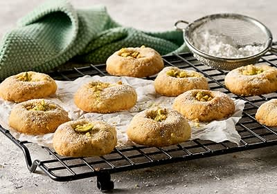 Elf runde Kekse mit Pistazien auf einem Gitterrost, daneben ein Sieb mit Puderzucker und ein grünes Handtuch