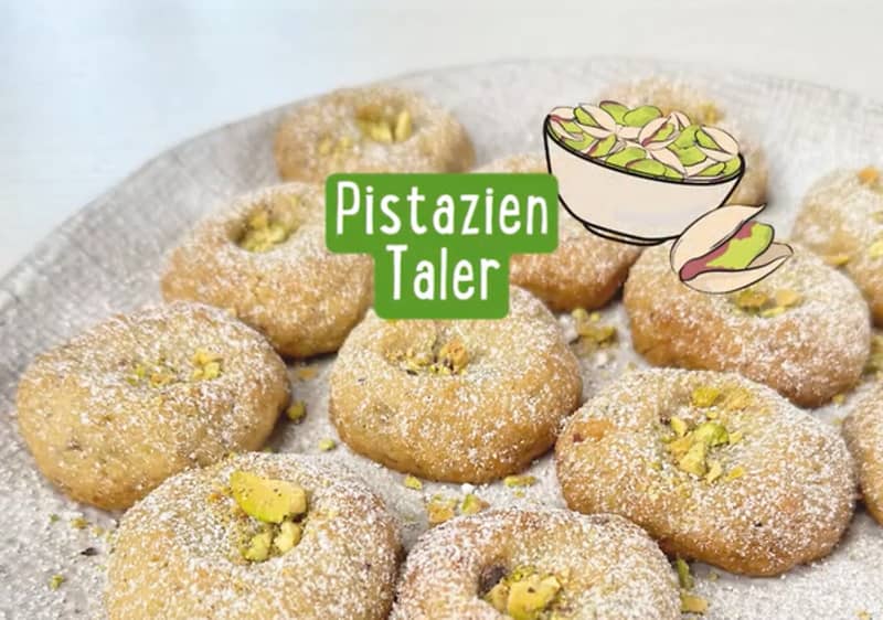 Mehrere Pistazien-Taler auf einem Teller, bestreut mit Puderzucker und gehackten Pistazien, daneben der Text „Pistazien Taler“ und eine Illustration einer Schale mit Pistazien