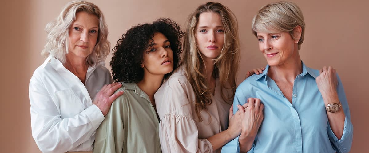 Vier Frauen mit unterschiedlichen Farbtypen stehen nebeneinander, tragen helle Kleidung und berühren sich sanft an den Schultern und Armen