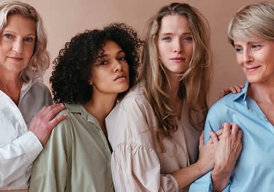 Vier Frauen mit unterschiedlichen Farbtypen stehen nebeneinander, tragen helle Kleidung und berühren sich sanft an den Schultern und Armen