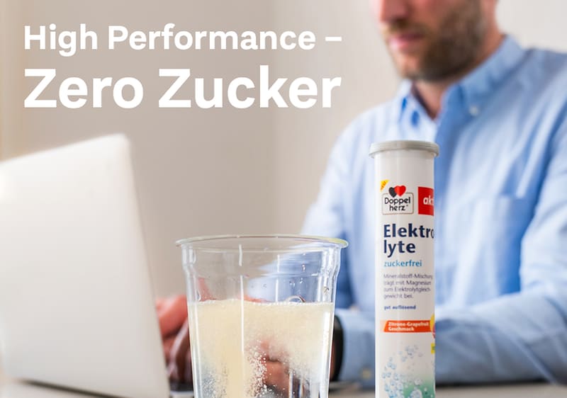 Ein Glas mit weißem Pulver, daneben eine Elektrolyte-Dose mit der Aufschrift „Zuckerfrei“, im Hintergrund der Text „High Performance – Zero Zucker“