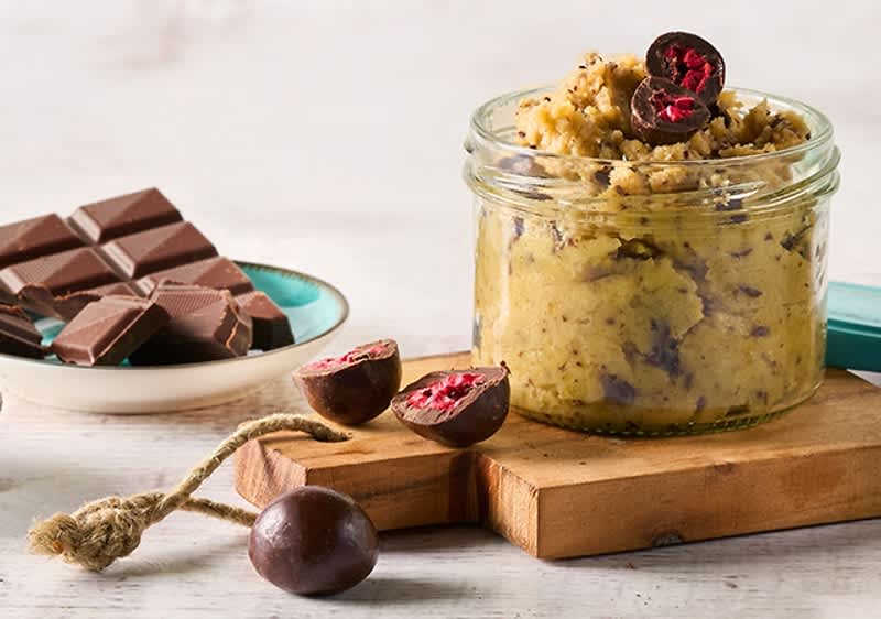 Ein Glas mit gelbem Peeling und dunklen Schokoladenstücken, daneben ein offenes Glas mit türkisfarbenem Deckel auf einem Holztisch
