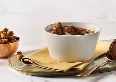 Ein weißer Keramikbecher mit Schokoladenmousse, dekoriert mit knusprigen Kugeln, steht auf einem gelben Stofftuch auf einem grauen Teller.