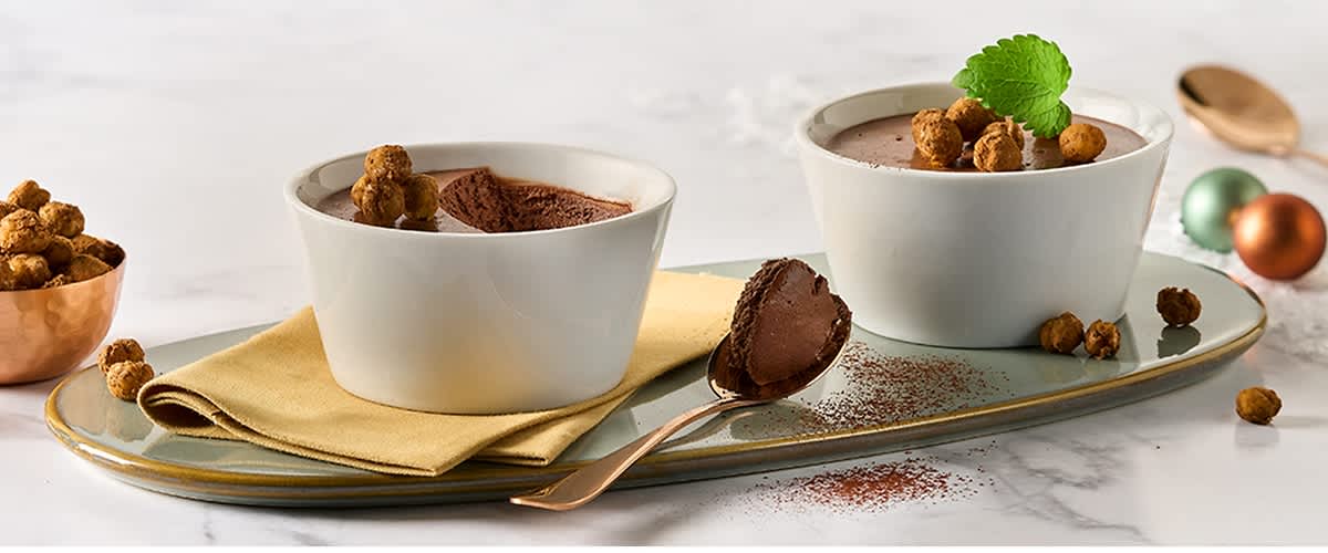 Zwei weiße Schalen mit Schokoladenmousse, dekoriert mit Keksen und Minzblatt, auf einem gelben Tuch, daneben ein Löffel mit Mousse