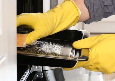 Eine Person mit gelben Gummihandschuhen reinigt ein schwarzes Backblech mit einem Schwamm in einem Ofen.