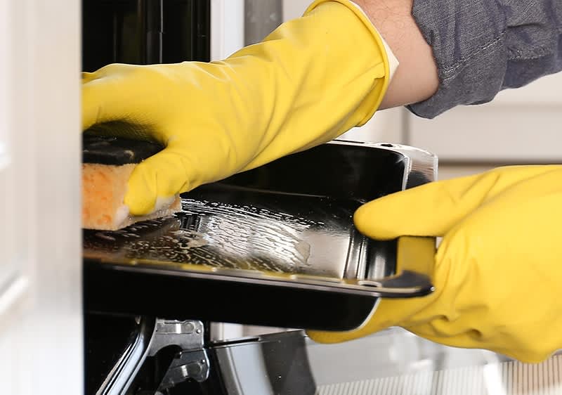 Eine Person mit gelben Gummihandschuhen reinigt ein schwarzes Backblech mit einem Schwamm in einem Ofen.
