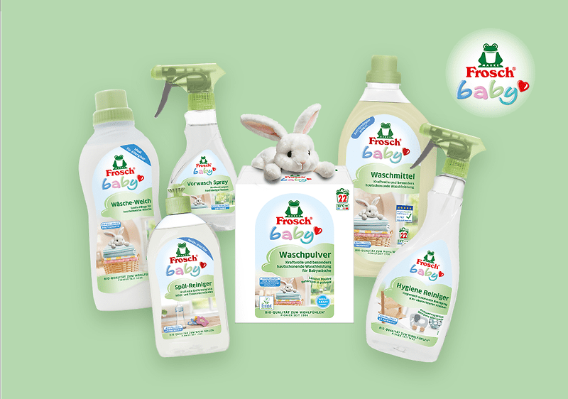 Sechs Frosch Baby Reinigungsprodukte auf pastellgrünem Hintergrund, mittig ein Waschpulver-Päckchen mit einem kleinen weißen Plüschhase, links und rechts Flüssigwaschmittel, Weichspüler, Spül-Reiniger und zwei Sprühflaschen mit sichtbaren Markenlogo und Produktbezeichnungen