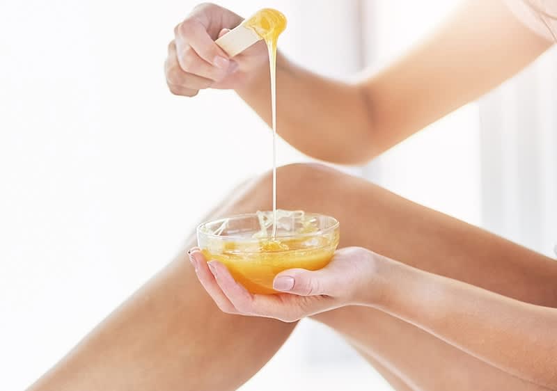 Eine Person hält eine Schale mit gelbem Wachs und trägt es mit einem Holzspatel auf die Haut auf.