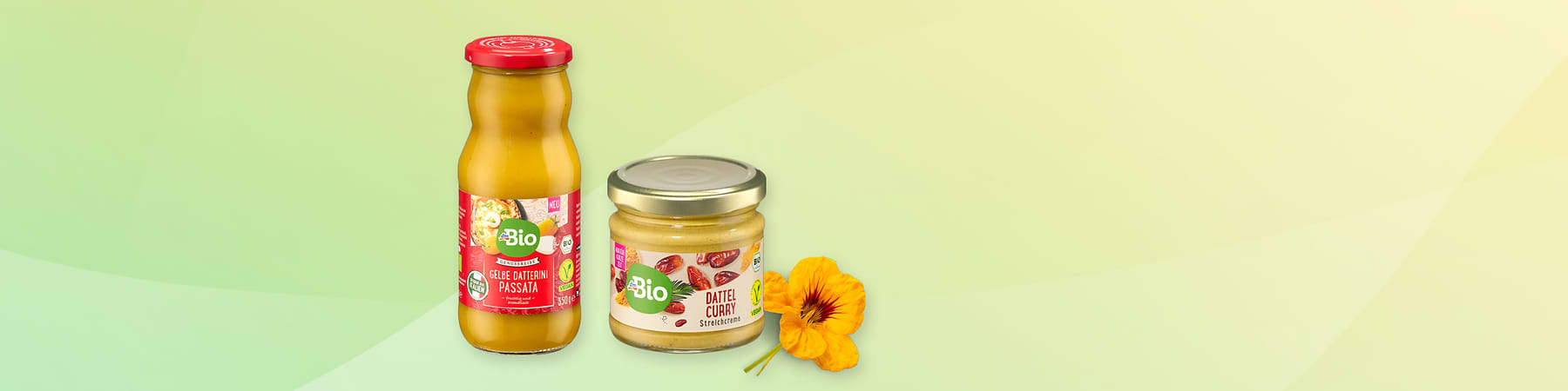 Zwei Bio-Produkte: links Gelee-Oliven-Passata in roter Flasche, rechts Dattel-Curry-Streichcreme im Glas. Daneben eine orange Blume.