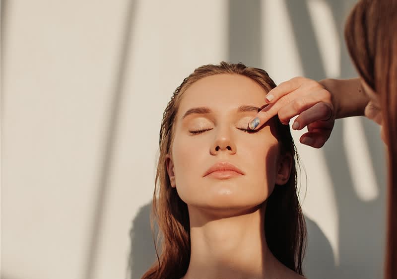 Eine Person mit langen Haaren wird von einer anderen Person frisiert, während Sonnenlicht durch ein Fenster fällt.