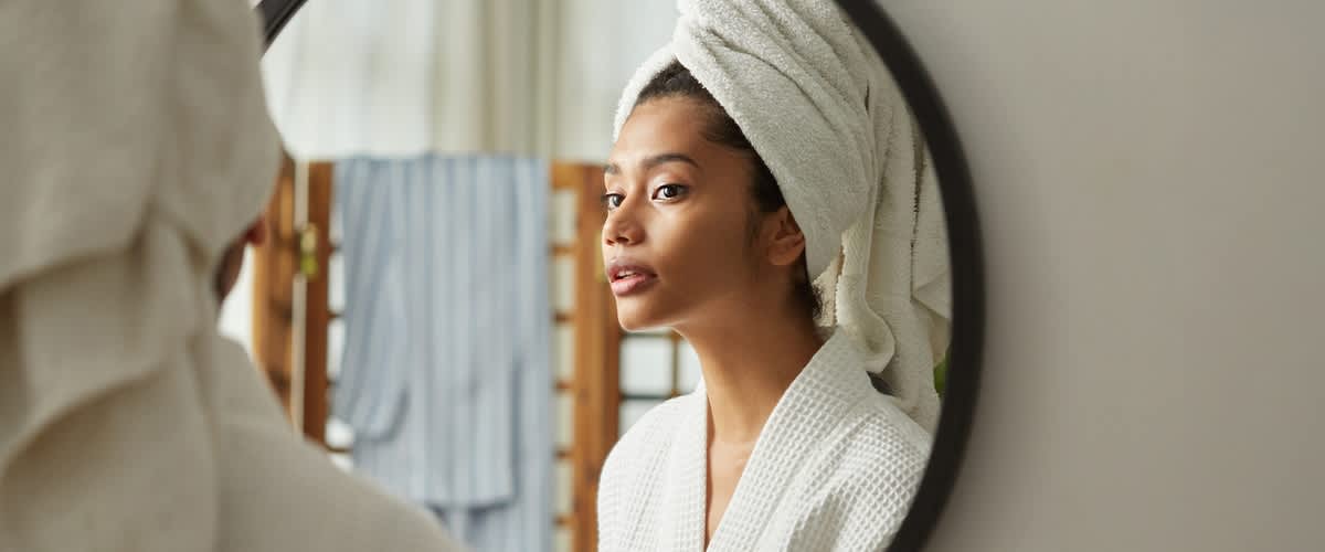 Eine Person in einem weißen Bademantel und mit einem Handtuch um den Kopf steht vor einem Spiegel. Im Hintergrund sind verschwommene Handtücher zu sehen.