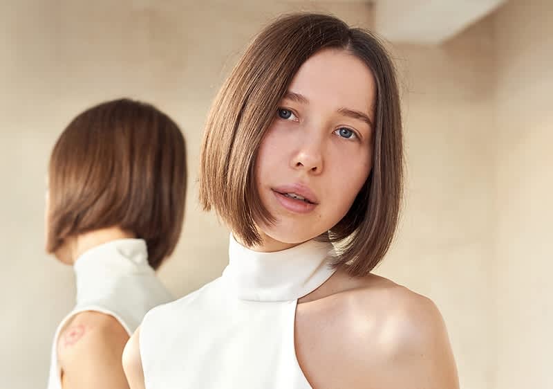 Eine Person mit einem asymmetrischen weißen Oberteil steht vor einem Spiegel, der ihren Rücken reflektiert.