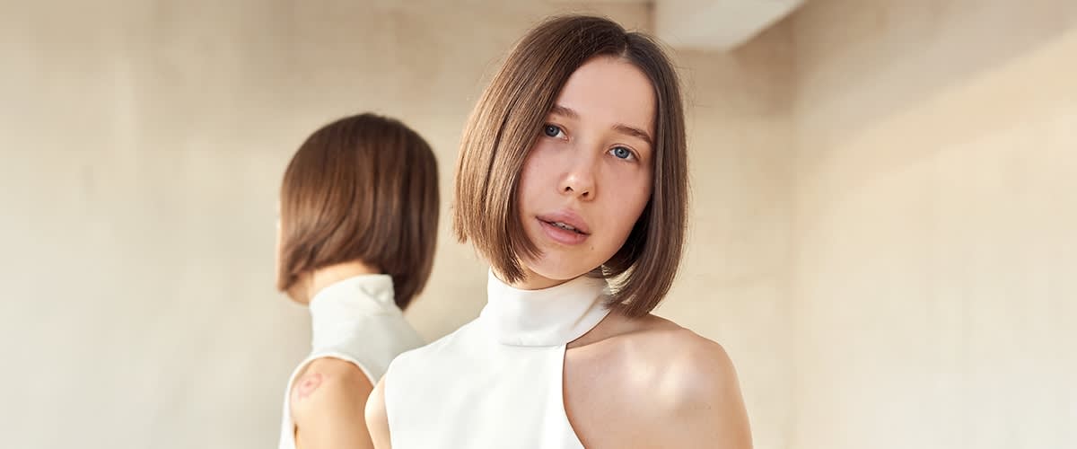 Eine Person mit schulterlangem, glattem Haar trägt ein weißes, ärmelloses Oberteil. Im Hintergrund ist ein Spiegel zu sehen.