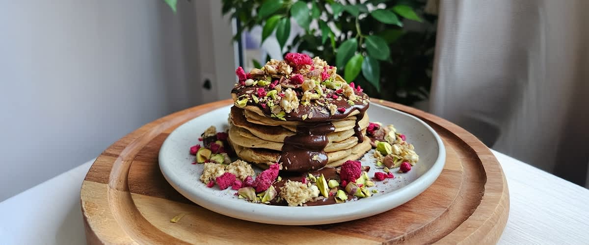 Ein Stapel Pfannkuchen mit Schokoladensauce, garniert mit Pistazien, Himbeeren und Haferflocken auf einem weißen Teller.