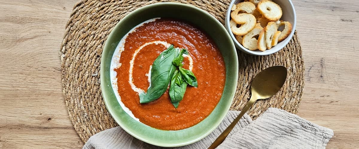 Tomatensuppe in einer grünen Schüssel, garniert mit Basilikumblättern und einem Sahnekringel, daneben eine Schale mit Brotchips.