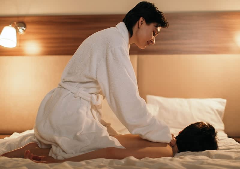 Eine Person in einem weißen Bademantel massiert eine liegende Person auf einem Bett in einem gemütlichen Raum mit warmem Licht.