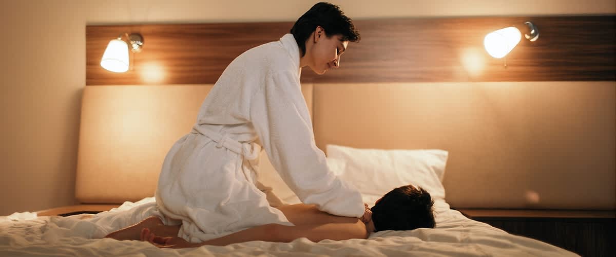 Eine Person in einem weißen Bademantel massiert eine liegende Person auf einem Bett in einem gemütlichen Raum mit warmem Licht.