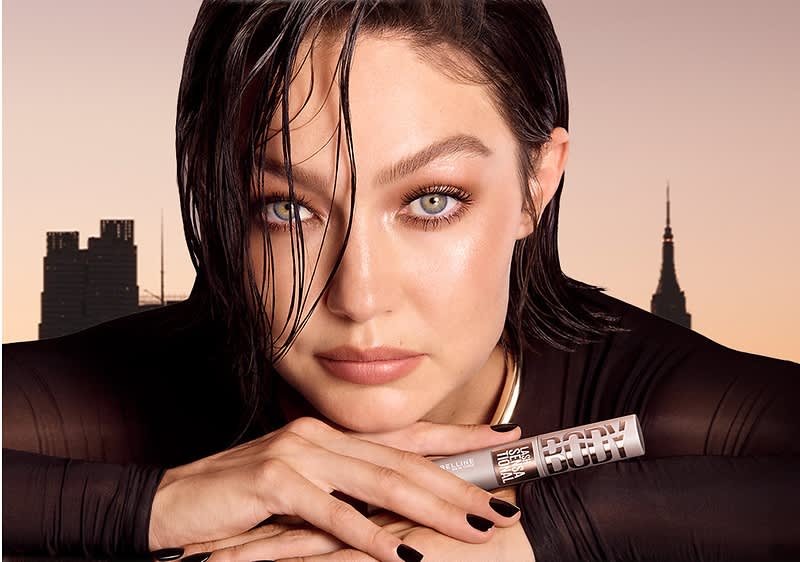 Eine Frau hält eine Tube mit der Aufschrift „Lash Sensational Body Mascara“ in der Hand. Der Hintergrund ist hellbraun.