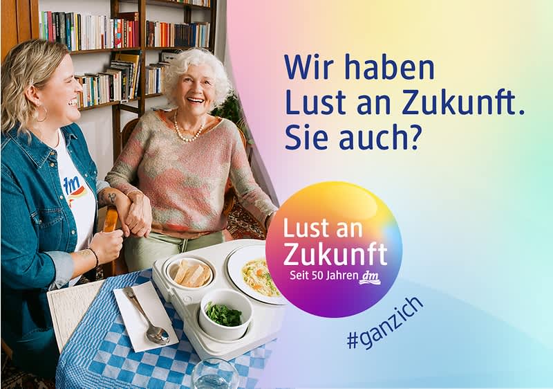 Zwei Personen sitzen an einem Tisch mit Essen. Links ein Regal mit Büchern. Text: „Wir haben Lust an Zukunft. Sie auch? Lust an Zukunft. Seit 50 Jahren dm #ganzich“