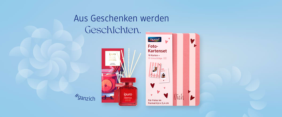 Hellblauer Hintergrund mit dem Schriftzug „Aus Geschenken werden Geschichten“, links ein rotes Raumduft-Set der Marke ipuro mit Glasflakon und Duftstäbchen, rechts eine rosa-weiße Fotokartenbox mit Aufschrift „Foto-Karten“ und Herzen sowie dem Wort „Dich“