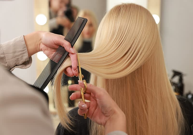 Friseurin kämmt und schneidet lange, glatte blonde Haare einer Kundin vor einem Spiegel