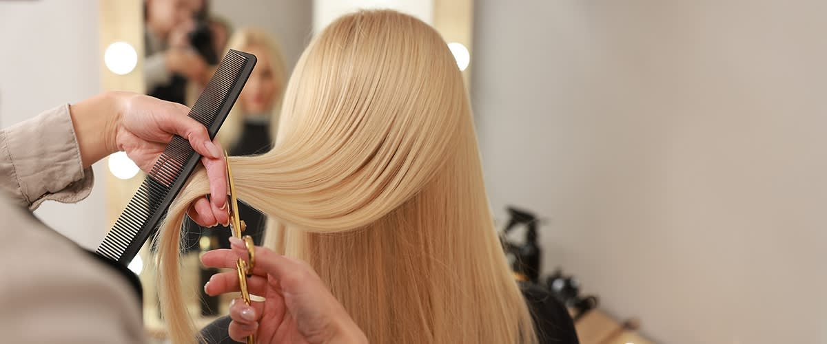 Friseurin kämmt und schneidet lange, glatte blonde Haare einer Kundin vor einem Spiegel