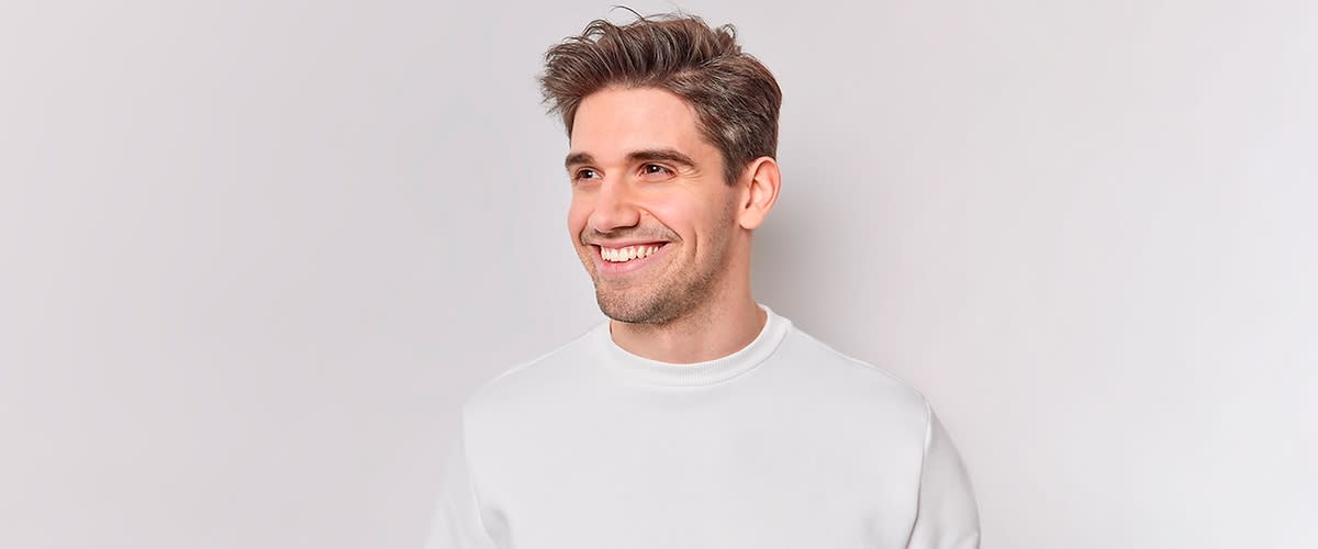 Junger Mann mit braunem, zurückgekämmt wirkendem Haar trägt ein weißes Sweatshirt vor hellem Hintergrund