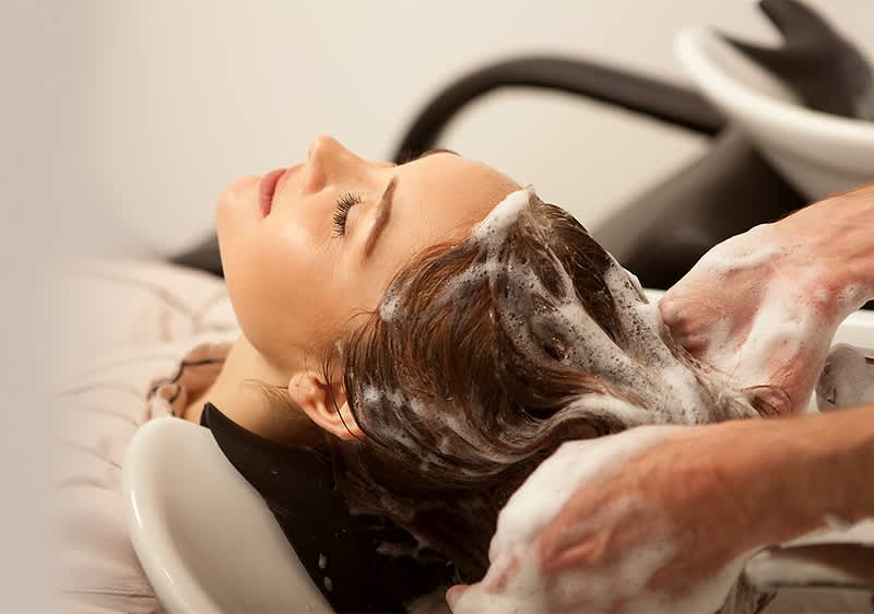 Person liegt mit nach hinten geneigtem Kopf in einem Friseurschwanenhals-Becken, Friseur wäscht schäumend die nassen Haare der Person, Hände mit Seifenschaum zu sehen