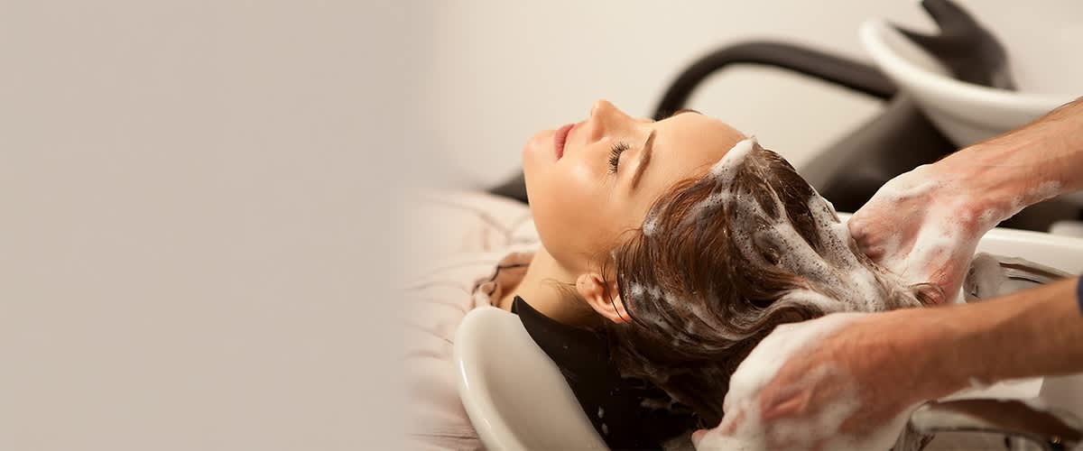 Person liegt mit nach hinten geneigtem Kopf in einem Friseurschwanenhals-Becken, Friseur wäscht schäumend die nassen Haare der Person, Hände mit Seifenschaum zu sehen