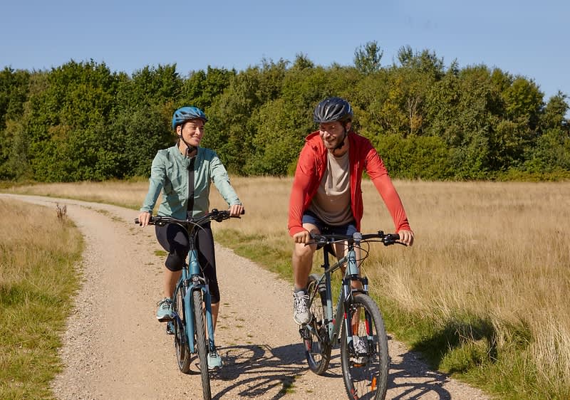 Zwei Personen fahren mit Fahrrädern auf einem Kiesweg durch eine Wiesenlandschaft, beide tragen Helme und Freizeitkleidung.