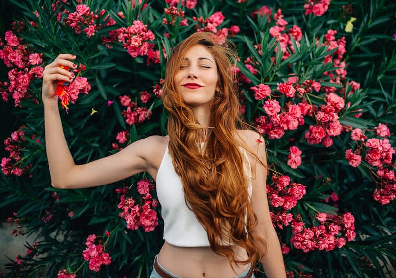 Junge Frau mit langem roten Haar in weißem Top liegt vor dichtem Busch mit zahlreichen pinken Blüten und hält eine einzelne Blüte in der rechten Hand
