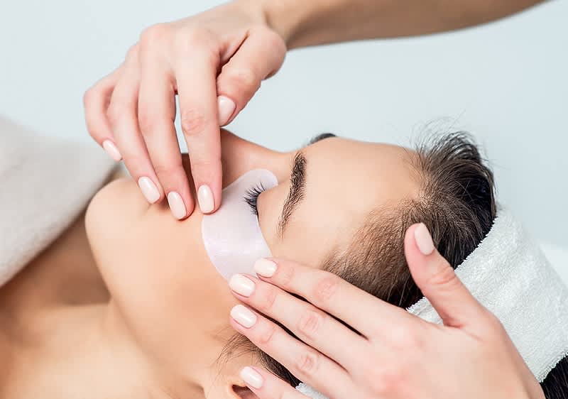 Frau mit Augenpads unter den unteren Augenlidern erhält von Händen eine kosmetische Behandlung am Gesicht, Stirnband hält Haare zurück