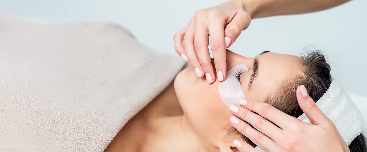 Frau mit Augenpads unter den unteren Augenlidern erhält von Händen eine kosmetische Behandlung am Gesicht, Stirnband hält Haare zurück