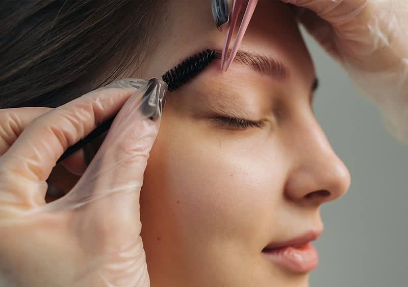 Junge Frau mit geschlossenen Augen, während eine Kosmetikerin per Pinzette und Bürstchen die Augenbraue formt