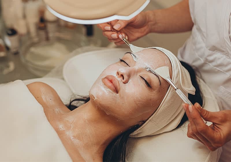 Nahaufnahme einer entspannenden Gesichtsbehandlung in einem Kosmetikstudio, eine Kosmetikerin führt mit einem Pinsel eine Maske auf das Gesicht einer liegenden Person auf, die Augen sind mit einem Stirnband bedeckt