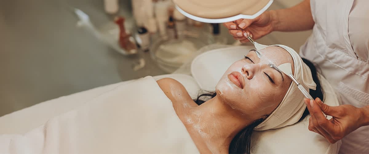 Nahaufnahme einer entspannenden Gesichtsbehandlung in einem Kosmetikstudio, eine Kosmetikerin führt mit einem Pinsel eine Maske auf das Gesicht einer liegenden Person auf, die Augen sind mit einem Stirnband bedeckt