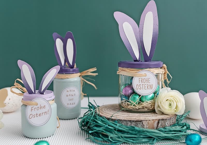 Drei dekorierte Gläser mit lila Hasenohren und Ostergrüßen, umgeben von bunten Eiern und einer weißen Blume auf einem Holztisch.
