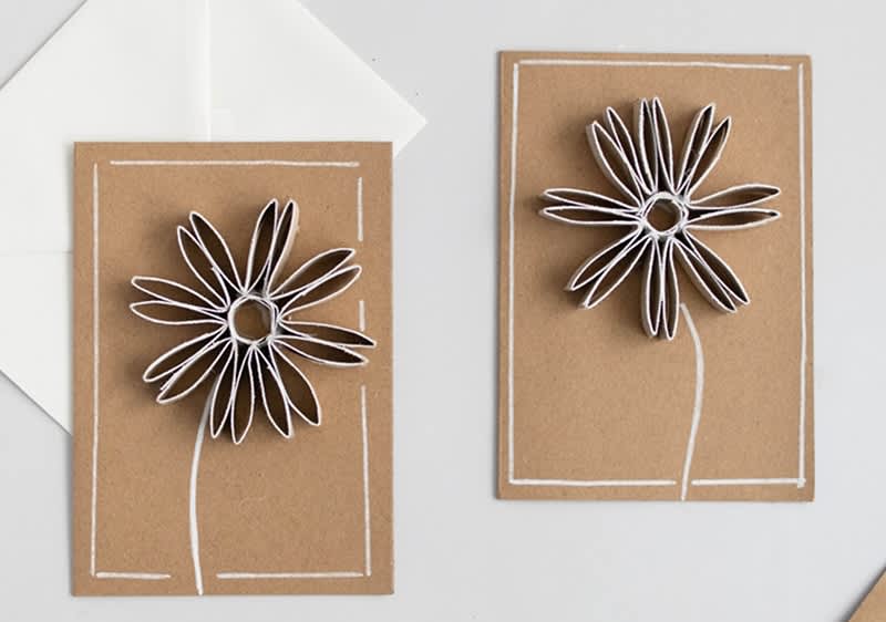 Zwei braune Karten mit weißen Blumen aus Papierstreifen, umrahmt von weißen Linien, liegen auf einem weißen Untergrund.