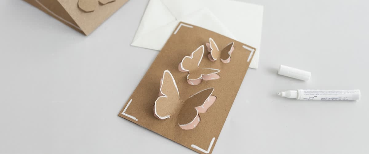 Karte aus braunem Papier mit ausgestanzten Schmetterlingen, daneben ein weißer Umschlag und ein weißer Stift