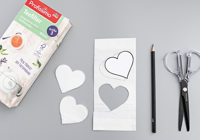 Eine Packung Profissimo Teefilter, ein Bleistift, eine Schere und ausgeschnittene Herzformen aus Teefilterpapier auf grauem Hintergrund